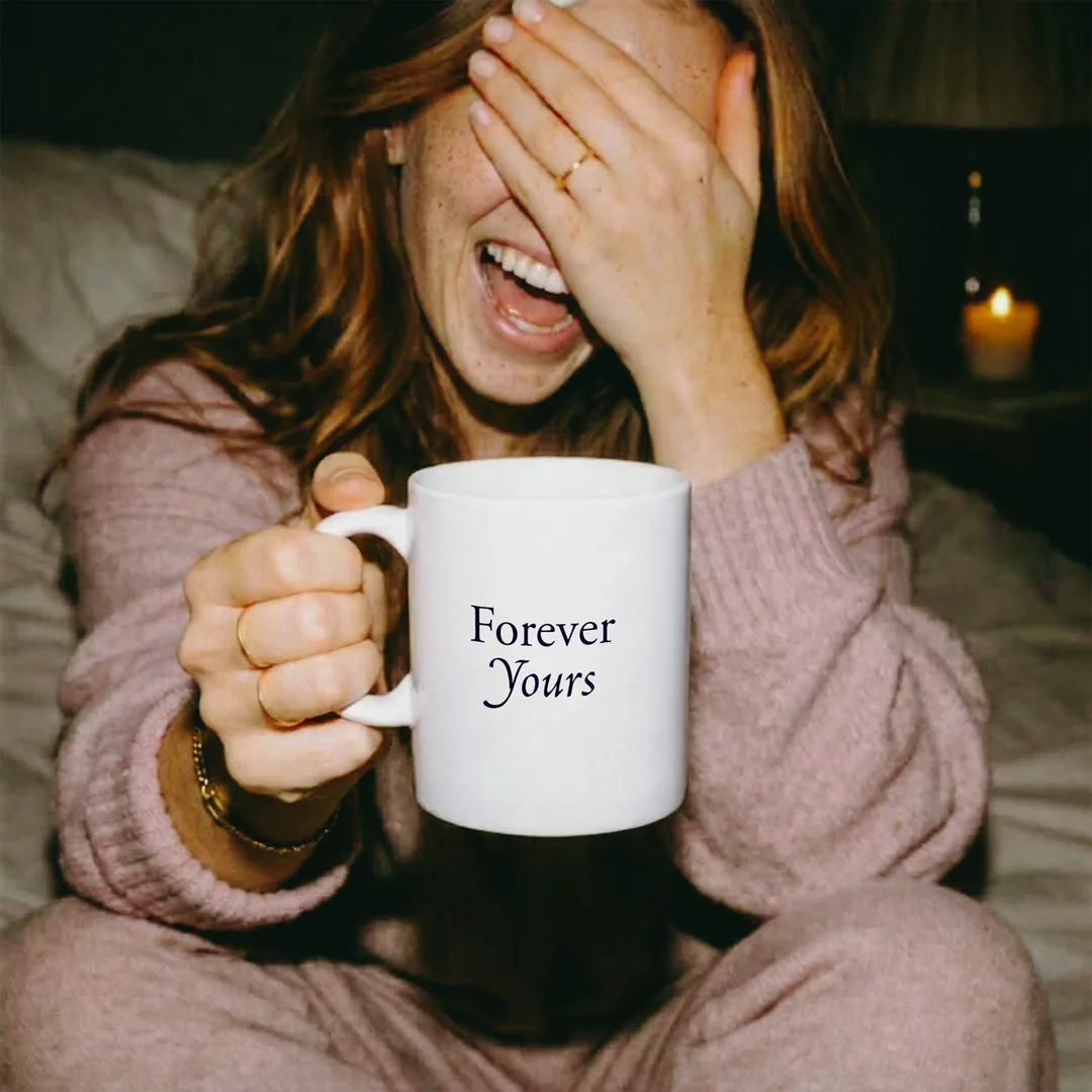 Person im rosa Strickpullover lacht hinter vorgehaltener Hand und hält die Forever Yours Tasse, dahinter Kerzenlicht