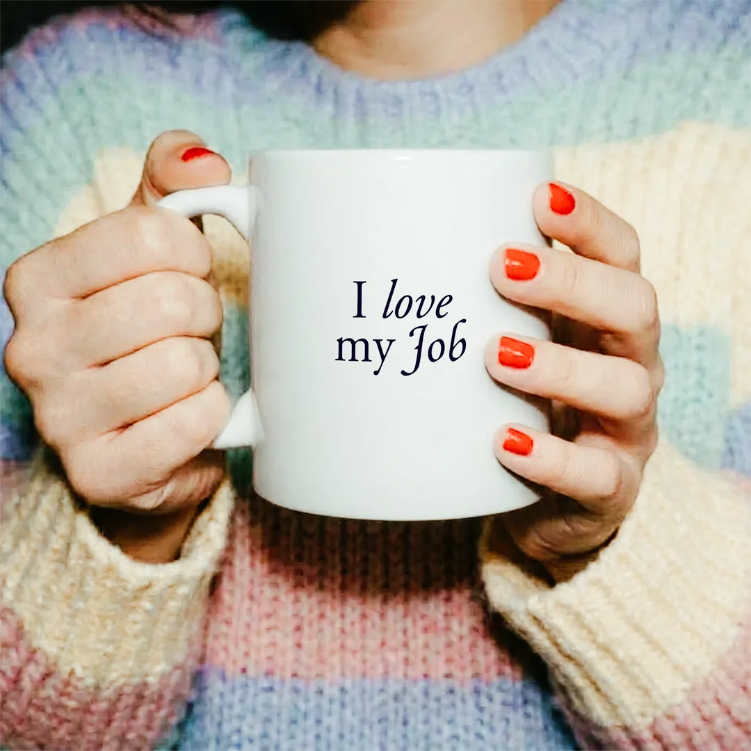 Zwei Hände mit rot lackierten Nägeln halten die I love my Job Tasse vor einem Pastell-Pullover