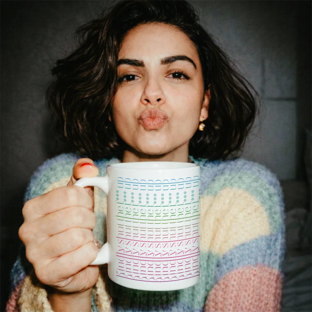 Person in buntem Strickpullover wirft einen Luftkuss, in der Hand die kindly shut the f*** up Tasse mit bunter Schrift