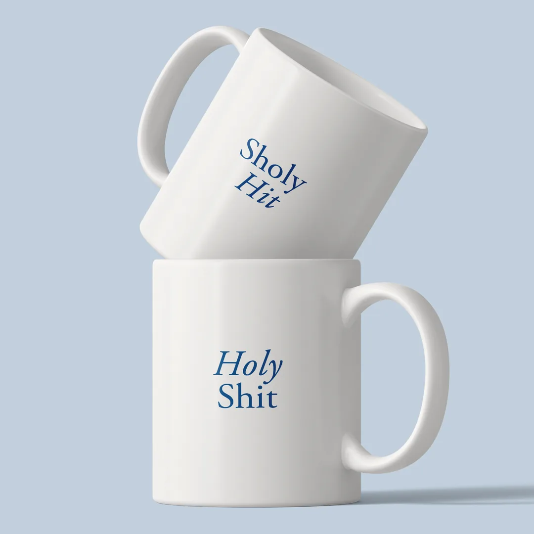 Two stacked Sholy Hit mugs on a light blue background, Sholy Hit on top, Holy Shit below