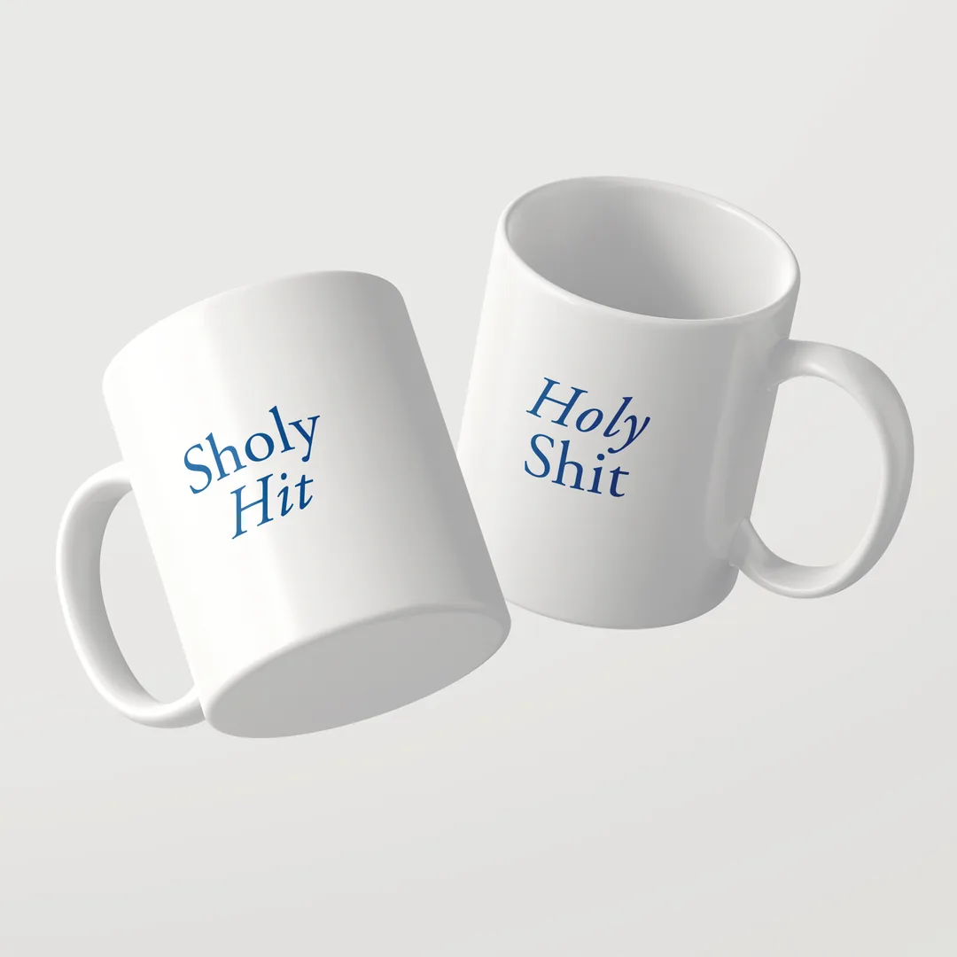 Two white Sholy Hit mugs by Studio SaySay, one reading Sholy Hit, the other Holy Shit in blue